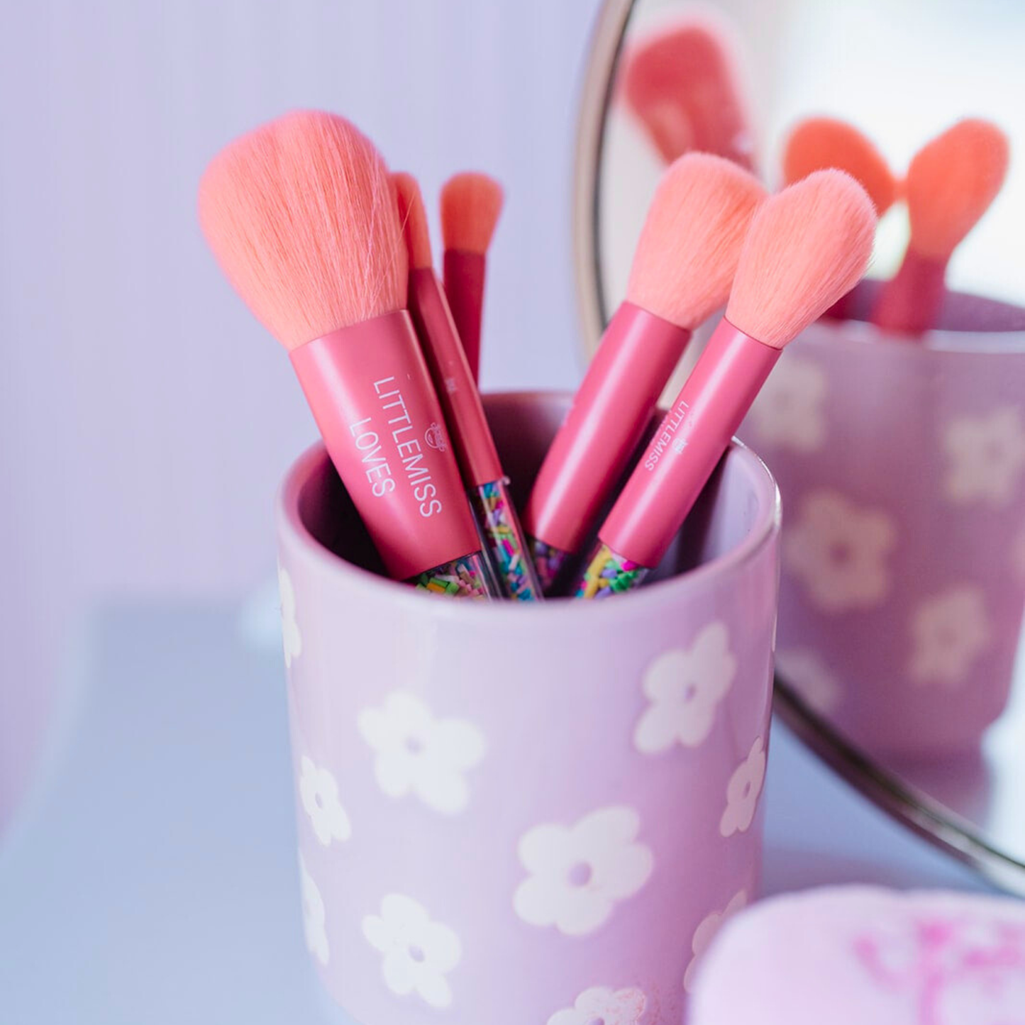 Makeup brushes with pink bristles in a purple cup with floral patterns on a light purple background.