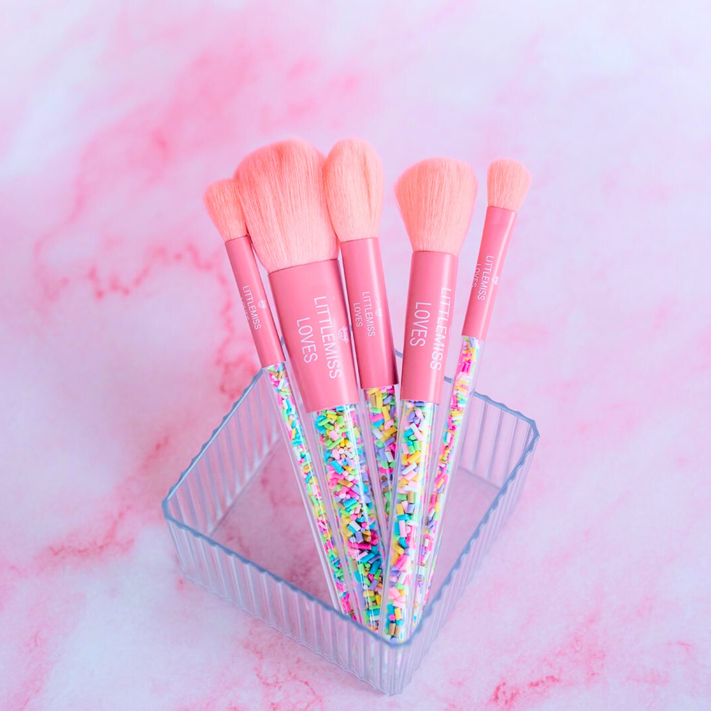 Set of pink makeup brushes with colorful handles in a clear container on a pink marble background