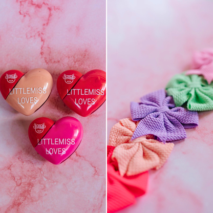 Heart-shaped containers with 'LITTLEMISS LOVES' branding on a pink marble background, accompanied by colorful hair ties.